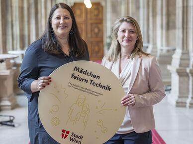 Vizebürgermeisterin und Frauenstadträtin Kathrin Gaál und Vizebürgermeisterin und Bildungsstadträtin Bettina Emmerling präsentieren die „Mädchen feiern Technik“-Workshops mit einem runden Schild, das von beiden gemeinsam gehaltne wird. Das Bild ist in den Arkadengängen des Rathauses aufgenommen worden. Kathrin Gaál trägt ein dunkelblaues Kleid, Bettina Emmerling eine dunkelblaue Hoseein beiges Oberteil mit Schalkragen und einen blassrosa Blazer darüber. Beide tragen ihre Haare offen.
