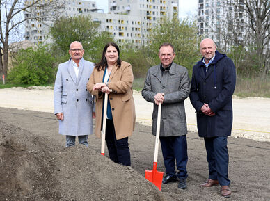 Dieter Groschopf, stellvertretender Geschäftsführer des wohnfonds_wien, Vizebürgermeisterin und Wohnbaustadträtin Kathrin Gaál, Bezirksvorsteher Gerald Bischof und WIGEBA-Vorstand Paul Steurer stehen an Spaten lehnend bei einem Sandhaufen. Die Aufnahme wurden offenbar zu einer kälteren Jahreszeit gemacht, da alle Protagonisten mit warmer Kleidung zu sehen sind.