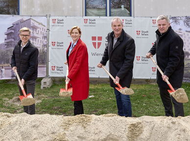 4 Personen stehen mit Spaten mit Sand darauf hinter einem Sandhaufen und lächeln in die Kamera. I Hintergrund ist ein Bauzaun mit Transparenten auf denen Visualisierungen des Gemeindebau NEU und das Logo von Stadt Wien/Wiener Wohnen zu erkennen ist.