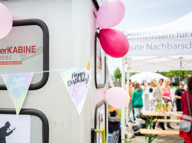 Links im Vordergrund ist eine Telefonzelle zu erkennen, die zu einer Bücherkabine umgebaut wurde. Sie ist mit Luftballons und mit einer Wimpel-Girlande geschmückt, auf der Happy Birthday steht. Im Hintergrund sind Menschen bei und unter einem wohnpartner-Pavillionzelt zu erkennen. auf einem Heurigentisch stehen Saftboxen. 