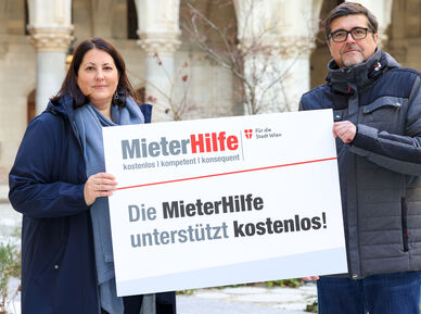 Kathrin Gaál und Christian Bartok stehen im Innenhof des Rathauses und halten gemeinsam eine Tafel auf der das Logo der MieterHilfe abgebildet ist und folgender Spruch zu lesen ist: "Die MieterHilfe unterstützt kostenlos!". Die beiden sind winterlich gekleidet.