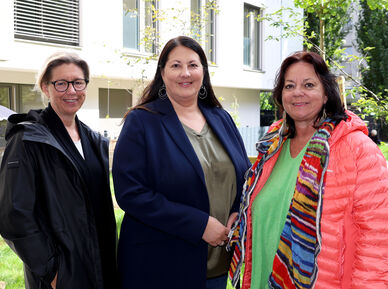 Wiener Wohnen Direktorin Karin Ramser, Vizebürgermeisterin und Wohnbaustadträtin Kathrin Gaál und wohnpartner-Bereichsleiterin Claudia Huemer stehen im begrünten Innenhof des neuen Gemeindebaus. Die drei sind trotz des offensichtlich freundlichen Wetters herbstlich gekleidet und lächeln freundlich in die Kamera.