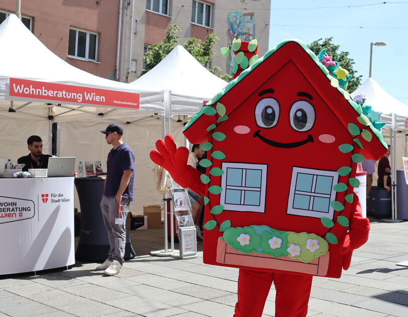 Das wienwohntbesser-Maskottchen Wohna Lisa, ein rotes Haus mit Armen und Beinen, einem Satteldach, einem freundlichen Lächeln im Gesicht und mit ausreichend Blumenschmuck steht auf einem Platz und winkt in die Kamera. Im Hintergrund ist ein Pagodenzelt sichtbar in dem an einem Pult mit Laptops darauf, zwei Personen eine weitere Person beraten, die davor steht.