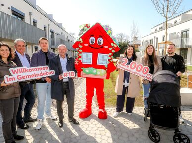 Sieben Personen stehen im Innenhof einer Wohnhausanlage. Die Anlage ist neu und hat drei Geschoße. In der Mitte der Gruppe steht das Maskottchen des Wiener Wohnbaus, Wohna Lisa. Sie ist ein rotes freundlich dreinblickendes Haus mit Satteldach auf zwei Beinen. Die Personen halten die Schriftzüge "Willkommen im Gemeindebau", "#18" und "2.000ste Gemeindewohnung NEU" in der Hand. Vor zwei Personen steht ein Kinderwagen. 