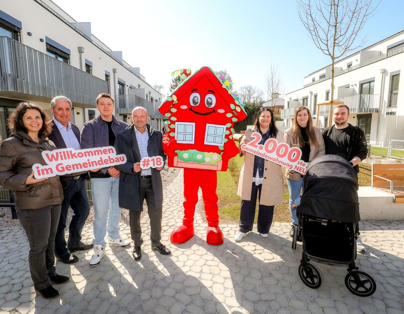 Sieben Personen stehen im Innenhof einer Wohnhausanlage. Die Anlage ist neu und hat drei Geschoße. In der Mitte der Gruppe steht das Maskottchen des Wiener Wohnbaus, Wohna Lisa. Sie ist ein rotes freundlich dreinblickendes Haus mit Satteldach auf zwei Beinen. Die Personen halten die Schriftzüge "Willkommen im Gemeindebau", "#18" und "2.000ste Gemeindewohnung NEU" in der Hand. Vor zwei Personen steht ein Kinderwagen. 