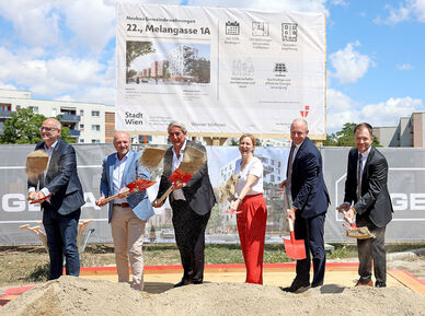 Sechs Personen mit Spaten stehen auf einer Baustelle vor einem großen Schild und schaufeln Erde in eine Sandkiste