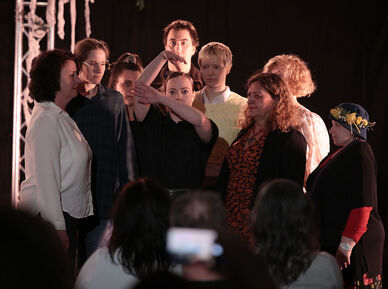 Mehrere Personen stehen knapp beisammen und blicken auf eine in der Mitte platzierte Person die mit ihren Armen die Äste eines Baumes darstellt. Der Hintergrund ist schwarz, im Vordergrund erkennt man Personen sitzend von hinten, die auf die Personengruppe schauen. 