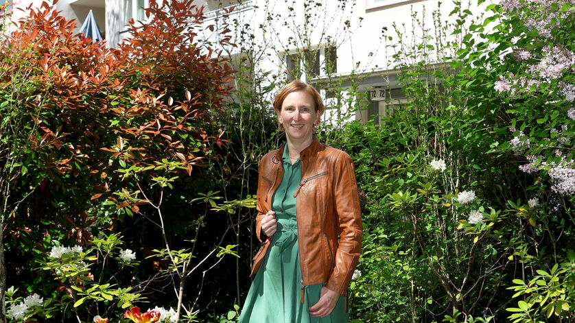 Frau in grünem Kleid und brauner Lederjacke steht in einem üppigen Garten vor einem mehrstöckigen Wohnhaus