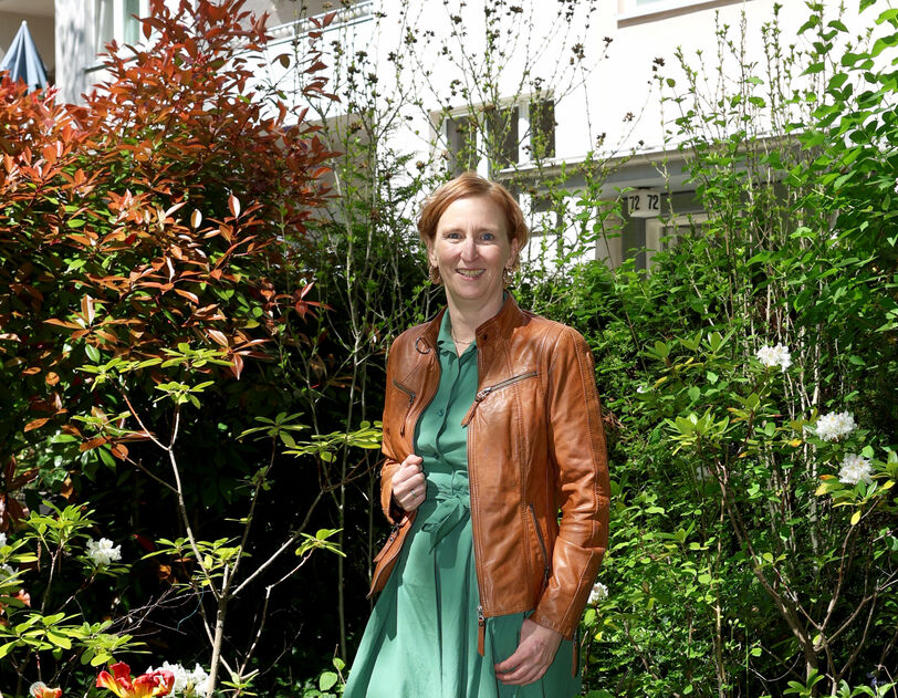 Frau in grünem Kleid und brauner Lederjacke steht in einem üppigen Garten vor einem mehrstöckigen Wohnhaus