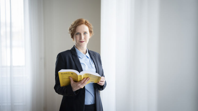 Frau mit roten Haaren in dunklem Blazer und hellblauer Bluse steht vor hellem Vorhang und hält ein aufgeschlagenes gelbes Buch, den Mietrechts-Kodex, in den Händen