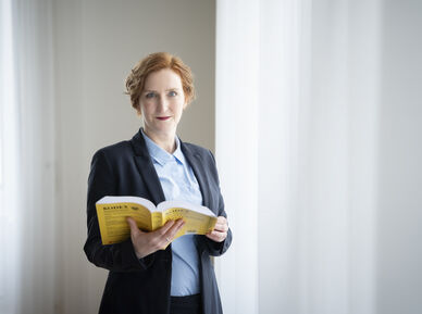 Frau mit roten Haaren in dunklem Blazer und hellblauer Bluse steht vor hellem Vorhang und hält ein aufgeschlagenes gelbes Buch, den Mietrechts-Kodex, in den Händen