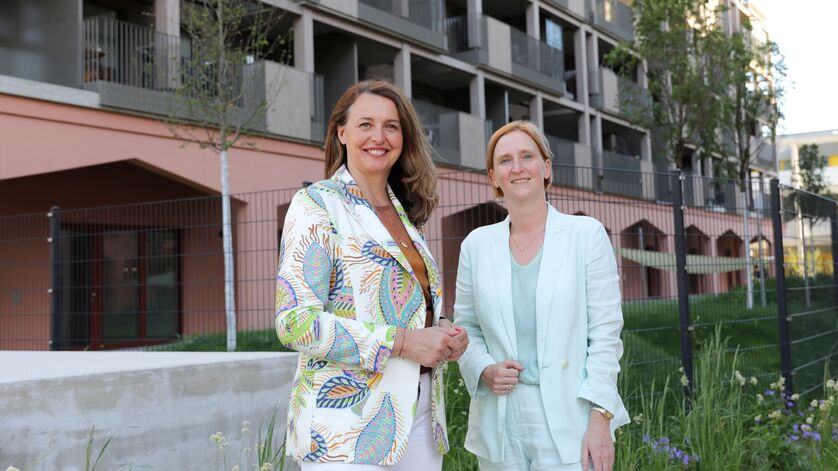 Zwei Frauen stehen vor einem modernen Wohngebäude, eine trägt einen gemusterten Blazer, die andere einen hellen Anzug