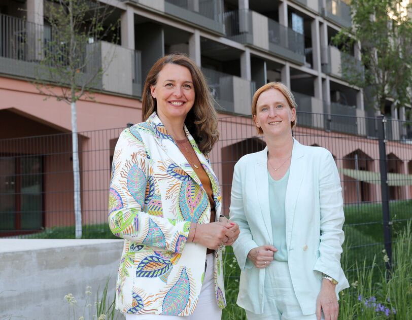 Zwei Frauen stehen vor einem modernen Wohngebäude, eine trägt einen gemusterten Blazer, die andere einen hellen Anzug