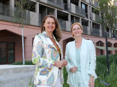 Zwei Frauen stehen vor einem modernen Wohngebäude, eine trägt einen gemusterten Blazer, die andere einen hellen Anzug