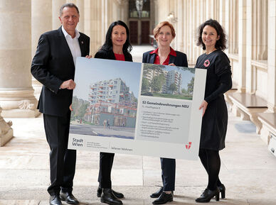 Drei weibliche und eine männliche Person stehen in den Arkaden des Wiener Rathauses und halten gemeinsam ein Schild auf dem Visualisierungen des Gemeindebau NEU zu sehen sind, sowie ein wenig Text