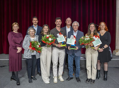 Gruppe von acht Personen vor rotem Vorhang und österreichischer Flagge, drei Personen halten Blumensträuße und Urkunden