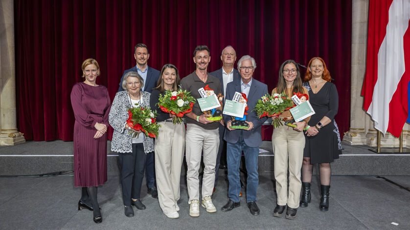 Gruppe von acht Personen vor rotem Vorhang und österreichischer Flagge, drei Personen halten Blumensträuße und Urkunden