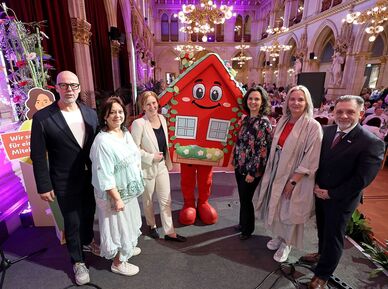 Gruppe von neun Personen steht in festlich beleuchtetem Saal neben wienwohntbesser-Maskottchen, ein rotes Haus mit Gesicht und Blumenschmuck, sie halten ein Schild mit Text 'Wir sorgen für ein gutes Miteinander'