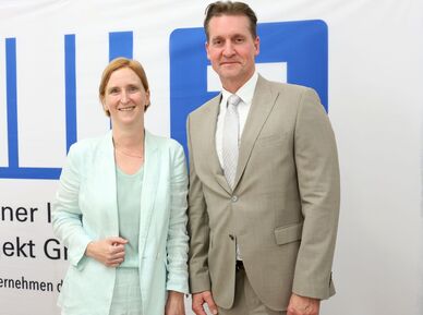 Zwei Personen in Anzügen stehen vor einem weißen Banner mit blauem Logo