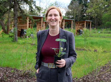 Person mit kurzem Haar und grauem Anzug hält einen kleinen Blumentopf mit einer Pflanze in einem Garten