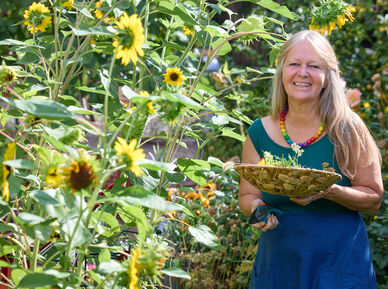 Frau mit langem Haar und bunter Halskette hält einen Korb mit Kräutern in einem Garten voller Sonnenblumen