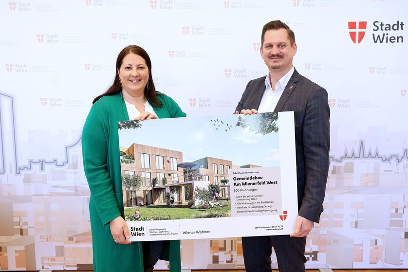 Eine Frau und ein Mann halten ein Schild mit der Visualisierung des neuen Gemeindebaus NEU in der Siedlung Wienerfeld West, erkennbar an der Beschriftung des Schildes. Sie lächeln in die Kamera.