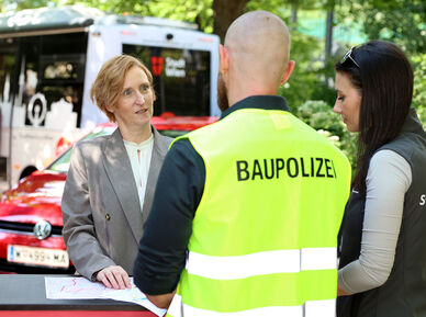 Drei Personen im Gespräch an einem Stehtisch, eine Frau trägt Blazer, ein Mann eine gelbe Weste mit Aufschrift "BAUPOLIZEI", eine weitere Frau trägt schwarze Weste