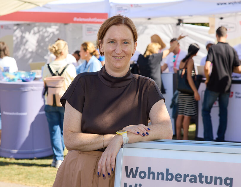 Frau lehnt an einem Schild mit der Aufschrift 'Wohnberatung Wien' auf einer Freiluftveranstaltung mit mehreren Menschen und Informationsständen im Hintergrund