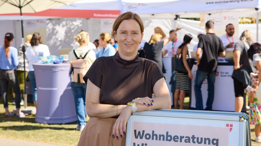 Frau lehnt an einem Schild mit der Aufschrift 'Wohnberatung Wien' auf einer Freiluftveranstaltung mit mehreren Menschen und Informationsständen im Hintergrund