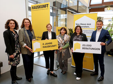 Fünf Personen stehen in einem modernen Innenraum und halten gelbe Schilder mit den Aufschriften '6 JAHRE HAUSKUNFT' und '23.000 BERATUNGEN', im Hintergrund gelbe Banner mit dem Schriftzug 'HAUSKUNFT'