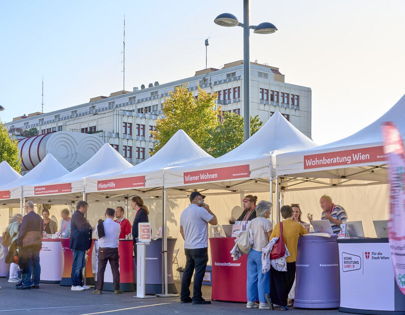 Menschen stehen an mehreren weißen Zelten mit roten Bannern und Informationsständen in einer sonnigen Straße vor einem großen Gebäude