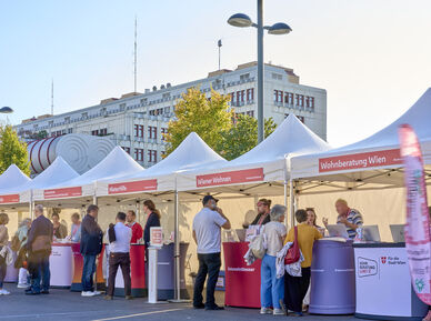 Menschen stehen an mehreren weißen Zelten mit roten Bannern und Informationsständen in einer sonnigen Straße vor einem großen Gebäude