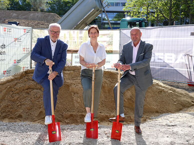 Drei Personen in formeller Kleidung stehen auf einer Baustelle und halten jeweils einen roten Spaten in der Hand, hinter ihnen ein großer Erdhaufen und Bauzäune mit dem Logo der Stadt Wien