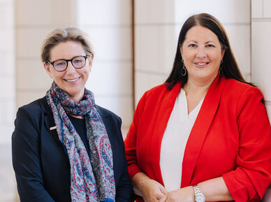 Zwei Frauen, Karin Ramser und Kathrin Gaál, stehen in einem Gang des Rathauses und lächeln in die Kamera. Beide sind businessmäßig mit Blazern gekleidet.