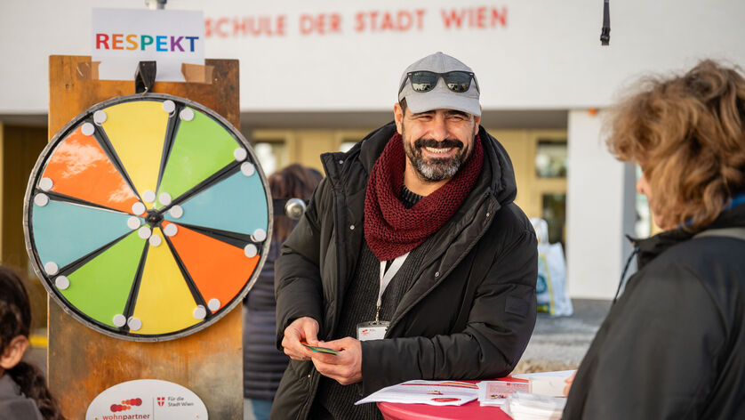 Ein freundlich lächelnder Mitarbeiter von wohnpartner, erkennbar an seinem Patch, steht neben einem Glücksrad . Er ist offensichtlich für kühleres Wetter gekleidet.