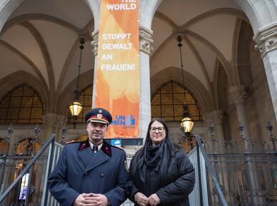 Ein hoher Polizeibeamter in Uniform mit Mantel und die Vizebürgermeisterin im Wintermantel posieren vor der am Wiener Rathaus gehissten orangen Flagge mit der Aufschrift "Orange the World" und "Stoppt Gewalt an Frauen!"