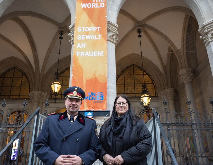 Ein hoher Polizeibeamter in Uniform mit Mantel und die Vizebürgermeisterin im Wintermantel posieren vor der am Wiener Rathaus gehissten orangen Flagge mit der Aufschrift "Orange the World" und "Stoppt Gewalt an Frauen!"